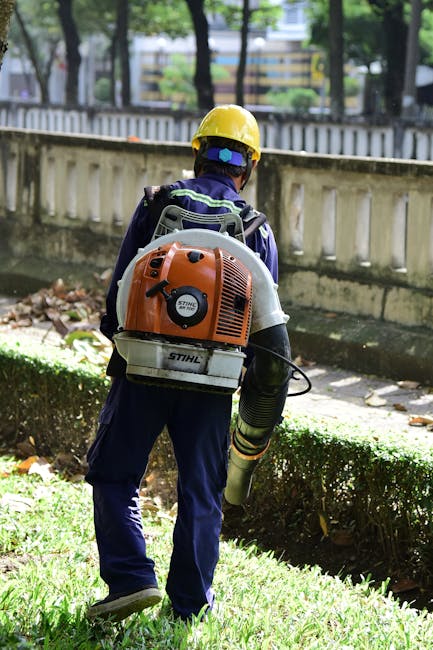 A gardener dressed in a blue uniform and yellow safety helmet using a leaf blower to clear fallen leaves and debris along a garden path. The garden features a neatly trimmed hedge, a grassy lawn, and